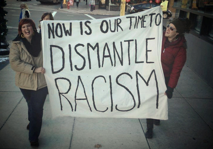 two people walking and holding a large sign that reads: now is our time to dismantle racism