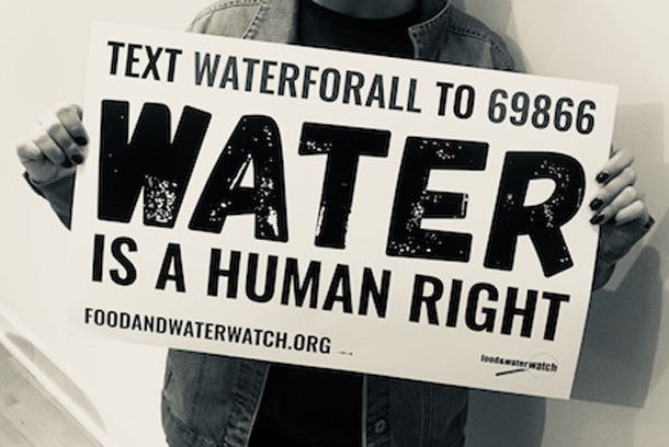 A person is holding a sign hat reads: Text WATERFORALL to 69866; water is a human right; foodandwaterwatch.org