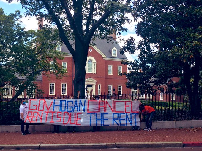 Two people hold a banner outside the Governor's mansion: Governor Hogan, Rent is Due. Cancel the Rent.