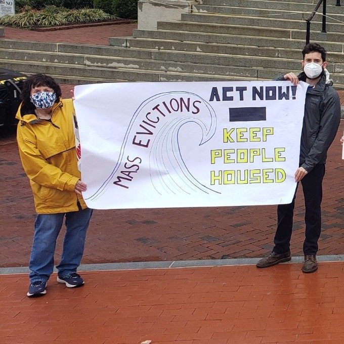 Jo and Matan hold a banner with a wave labeled Mass Evictions and text that says Act Now! Keep People Housed.