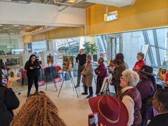 Molly Amster delivers remarks at University of Baltimore library beside paintings by Kimberly Sheridan